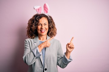 Middle age beautiful woman wearing bunny ears standing over isolated pink background smiling and looking at the camera pointing with two hands and fingers to the side.