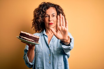 Middle age beautiful woman holding cake standing over isolated yellow background with open hand...