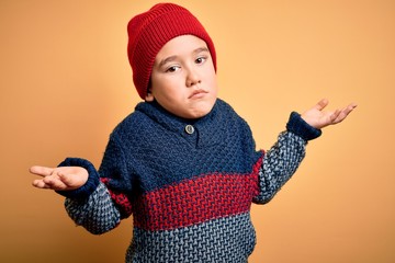 Young little boy kid wearing wool cap and winter sweater over yellow isolated background clueless and confused expression with arms and hands raised. Doubt concept.