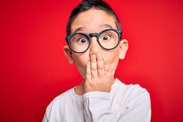 Young little smart boy kid wearing nerd glasses over red isolated background cover mouth with hand shocked with shame for mistake, expression of fear, scared in silence, secret concept © Krakenimages.com