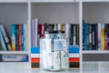 Glass jar full of 20 euro € banknotes money and books on the white table in front of the shelf savings and study scholarship concept wealth as knowledge investment