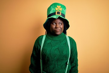 Plus size african american woman with braids wearing green hat with clover on st patricks day smiling looking to the side and staring away thinking.