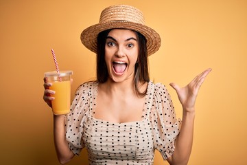 Young beautiful brunette woman on vacation wearing summer hat drinking orange juice very happy and excited, winner expression celebrating victory screaming with big smile and raised hands