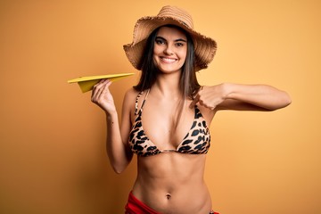 Young beautiful woman on vacation wearing bikini and summer hat holding paper airplane with surprise face pointing finger to himself