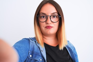 Beautiful woman wearing denim shirt and glasses make selfie over isolated white background with a confident expression on smart face thinking serious