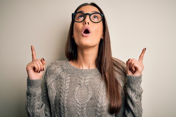 Young beautiful brunette woman wearing casual sweater and glasses over white background amazed and surprised looking up and pointing with fingers and raised arms.