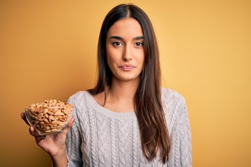 Young beautiful brunette woman holding bowl with peanuts over isolated yellow background with a confident expression on smart face thinking serious