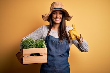 Young beautiful brunette gardener woman wearing apron and hat holding box with plants happy with...