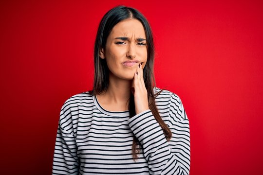 Young beautiful brunette woman wearing casual striped t-shirt over red background touching mouth with hand with painful expression because of toothache or dental illness on teeth. Dentist