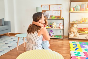 Young beautiful teacher and blond toddler girl hugging at kindergarten