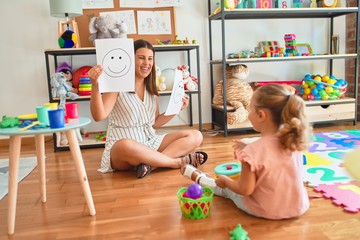 Beautiful psycologist and blond toddler girl sitting on the floor doing therapy using emoji...