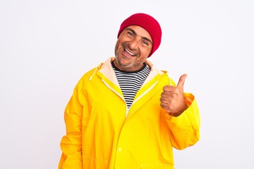 Middle age man wearing rain coat and woolen hat standing over isolated white background doing happy thumbs up gesture with hand. Approving expression looking at the camera showing success.