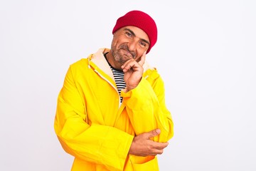 Middle age man wearing rain coat and woolen hat standing over isolated white background looking confident at the camera smiling with crossed arms and hand raised on chin. Thinking positive.