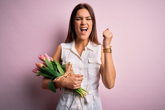 Young Beautiful Brunette Woman Holding Bouquet Of Tulips Flowers Over Pink Background Angry And Mad Raising Fist Frustrated And Furious While Shouting With Anger. Rage And Aggressive Concept.