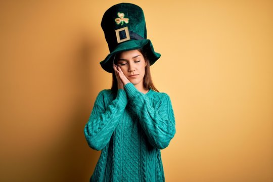 Young beautiful brunette woman wearing green hat with clover celebrating saint patricks day sleeping tired dreaming and posing with hands together while smiling with closed eyes.