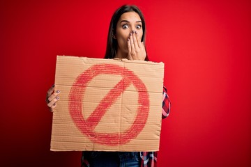 Young beautiful brunette woman holding banner with prohibited signal over red background cover...