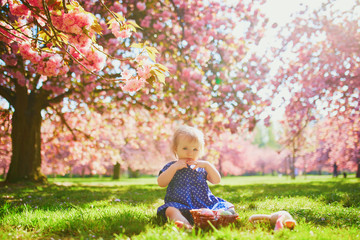 Cute one year old girl sitting on the grass and eating strawberries