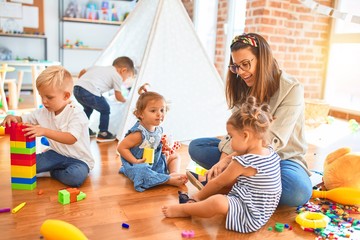 Beautiful teacher and group of toddlers playing around lots of toys at kindergarten - obrazy, fototapety, plakaty