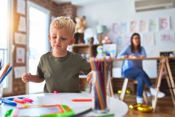 Fototapeta premium Young caucasian child playing at playschool with teacher. Mother and son at playroom drawing a draw with color pencils, young woman at the background sitting on desk.