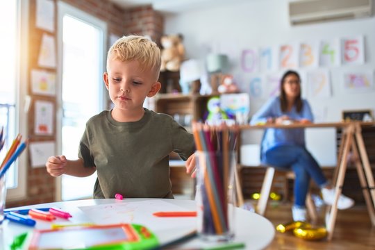 Young caucasian child playing at playschool with teacher. Mother and son at playroom drawing a draw with color pencils, young woman at the background sitting on desk.