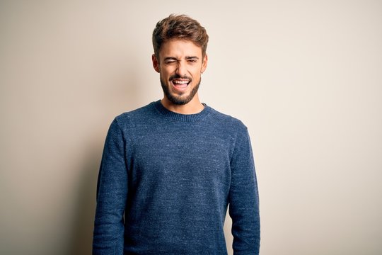 Young handsome man with beard wearing casual sweater standing over white background winking looking at the camera with sexy expression, cheerful and happy face.