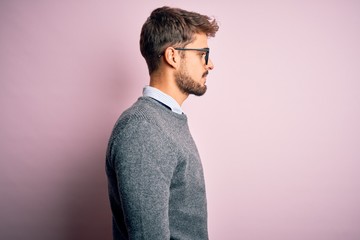 Young handsome man with beard wearing glasses and sweater standing over pink background looking to...
