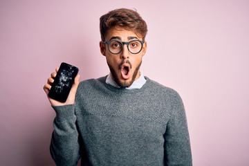 Young man with beard wearing glasses holding broken and craked smartphone scared in shock with a...
