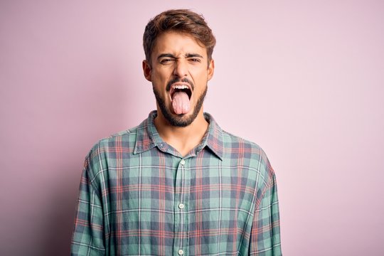 Young handsome man with beard wearing casual shirt standing over pink background sticking tongue out happy with funny expression. Emotion concept.