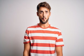 Young handsome man with beard wearing striped t-shirt standing over white background making fish face with lips, crazy and comical gesture. Funny expression.