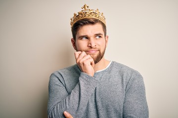 Young blond man with beard and blue eyes wearing golden crown of king with hand on chin thinking...