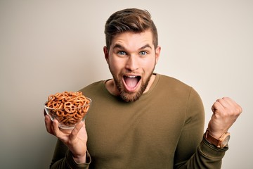 Young blond German man with beard and blue eyes holding bowl with baked pretzel screaming proud and celebrating victory and success very excited, cheering emotion