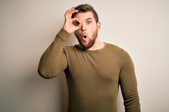 Young blond man with beard and blue eyes wearing green sweater over white background doing ok gesture shocked with surprised face, eye looking through fingers. Unbelieving expression.