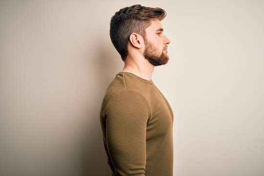 Young blond man with beard and blue eyes wearing green sweater over white background looking to side, relax profile pose with natural face with confident smile.