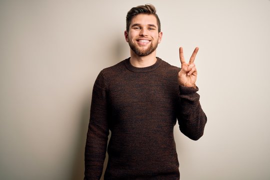 Young handsome blond man with beard and blue eyes wearing casual sweater standing smiling with happy face winking at the camera doing victory sign. Number two.