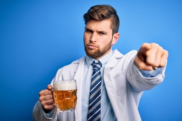Young blond doctor man with beard and blue eyes wearing coat drinking jar of beer pointing with finger to the camera and to you, hand sign, positive and confident gesture from the front