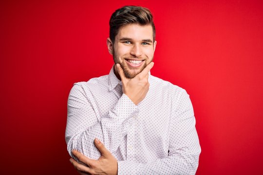 Young blond businessman with beard and blue eyes wearing elegant shirt over red background looking confident at the camera smiling with crossed arms and hand raised on chin. Thinking positive.