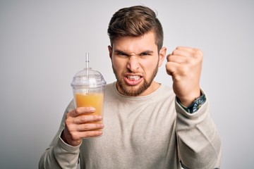 Young blond man with beard and blue eyes drinking healthy orange smoothie annoyed and frustrated shouting with anger, crazy and yelling with raised hand, anger concept