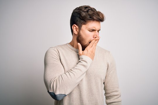 Young handsome blond man with beard and blue eyes wearing casual sweater bored yawning tired covering mouth with hand. Restless and sleepiness.
