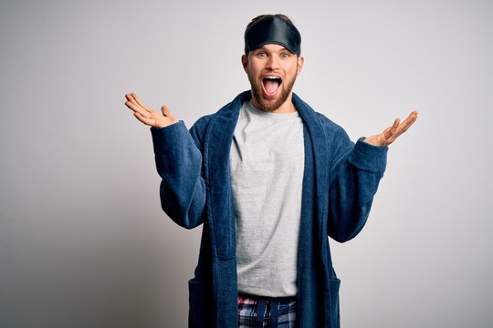 Young blond man with beard and blue eyes wearing sleep mask and pajama celebrating crazy and amazed for success with arms raised and open eyes screaming excited. Winner concept