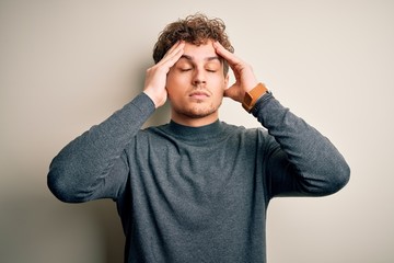 Young blond handsome man with curly hair wearing casual sweater over white background suffering from headache desperate and stressed because pain and migraine. Hands on head.