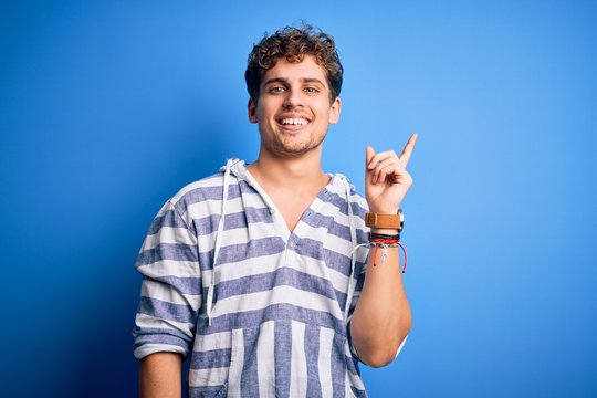 Young blond handsome man with curly hair wearing casual striped sweatshirt with a big smile on face, pointing with hand and finger to the side looking at the camera.