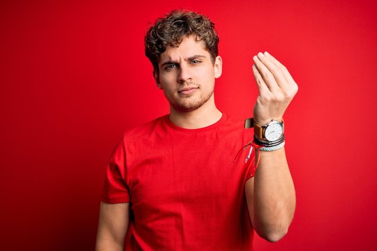 Young blond handsome man with curly hair wearing casual t-shirt over red background Doing Italian gesture with hand and fingers confident expression