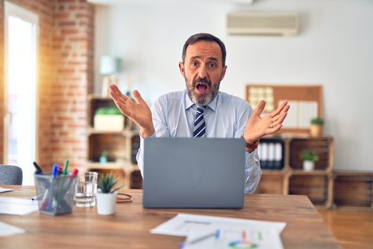 Middle age handsome businessman wearing tie sitting using laptop at the office clueless and confused expression with arms and hands raised. Doubt concept.