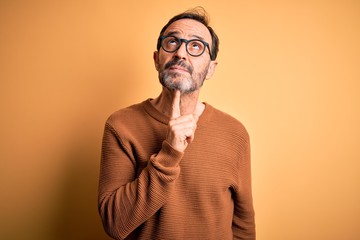 Middle age hoary man wearing brown sweater and glasses over isolated yellow background Thinking concentrated about doubt with finger on chin and looking up wondering