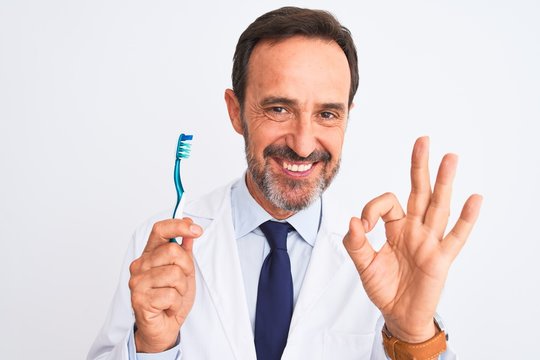 Middle age dentist man holding toothbrush standing over isolated white background doing ok sign with fingers, excellent symbol