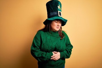 Young beautiful plus size woman wearing green hat with clover celebrating saint patricks day with hand on stomach because indigestion, painful illness feeling unwell. Ache concept.