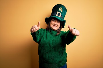 Young beautiful plus size woman wearing green hat with clover celebrating saint patricks day approving doing positive gesture with hand, thumbs up smiling and happy for success. Winner gesture.