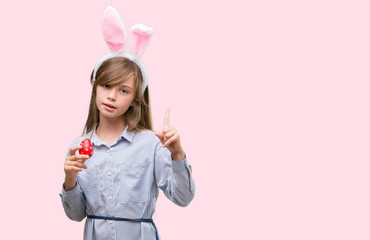 Young blonde child wearing easter bunny ears surprised with an idea or question pointing finger with happy face, number one