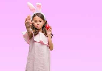 Obraz na płótnie Canvas Brunette hispanic girl wearing easter rabbit ears with open hand doing stop sign with serious and confident expression, defense gesture