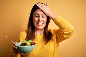 Young beautiful woman eating healthy fresh salad over yellow background stressed with hand on head, shocked with shame and surprise face, angry and frustrated. Fear and upset for mistake.